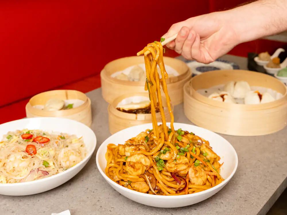 Shrimp noodles lifted from the bowl with chopsticks, with steamers of buns and dumplings behind